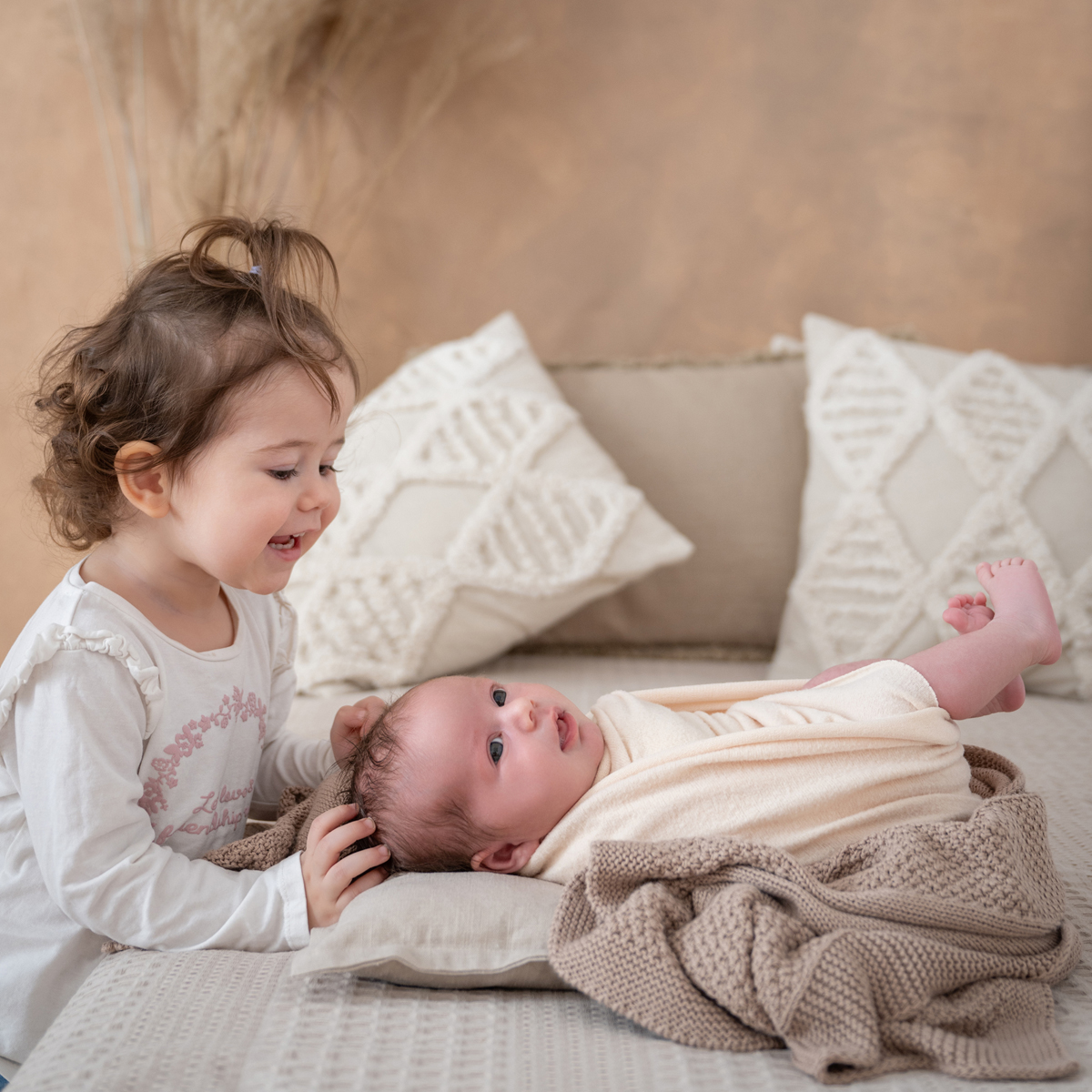 Kleines Mädchen beugt sich lachend über ihr eingewickeltes Babygeschwisterchen, das aufmerksam auf dem Bett liegt.